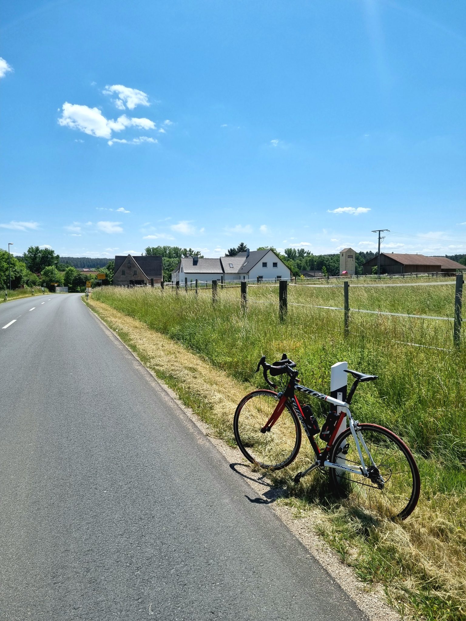 A red, white and black racing bike leaning at the side of the road in front of a rural village on a sunny day.
