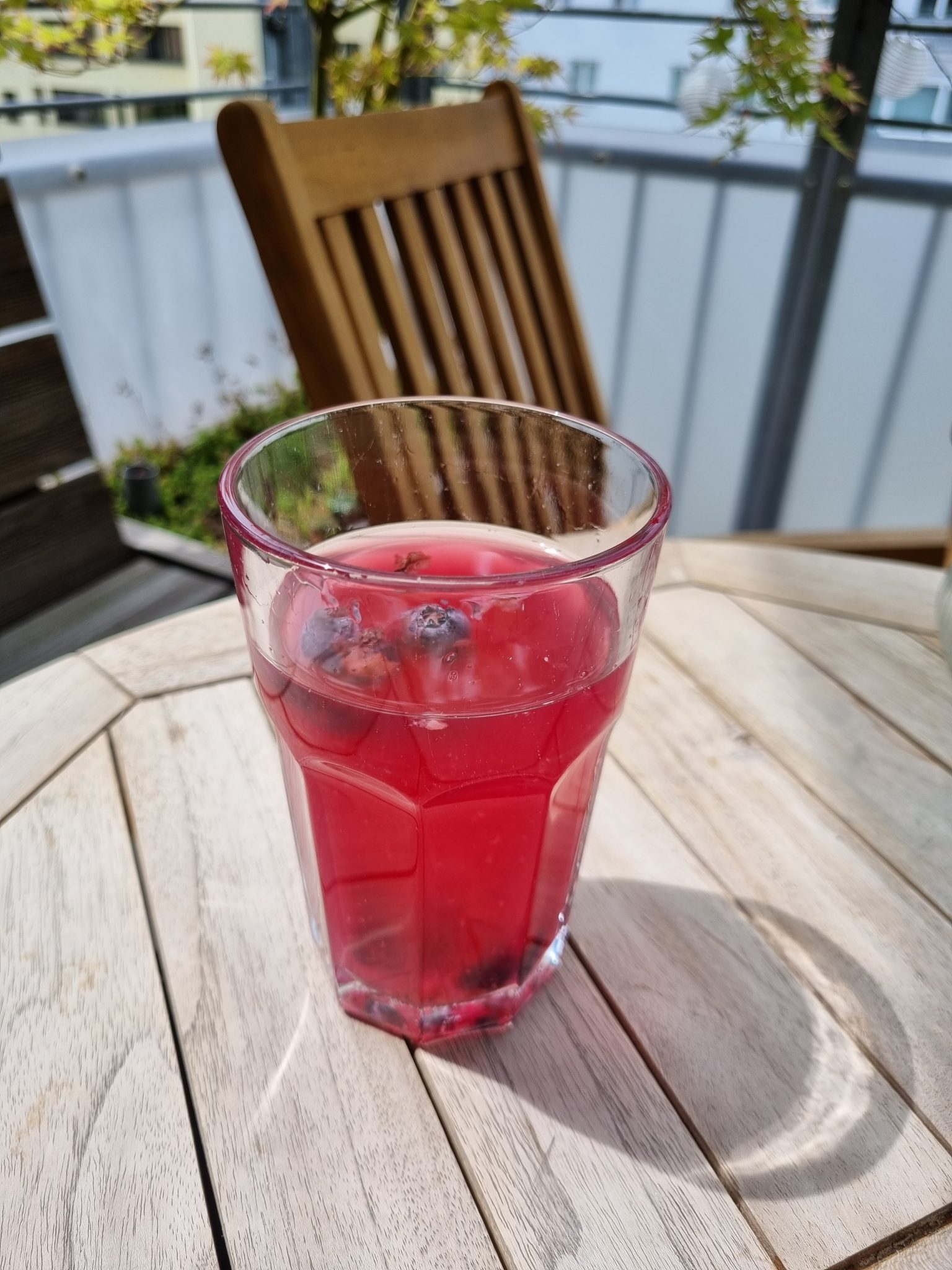 A glass full with bright magenta homebrew kombucha with blueberries in it standing on a wooden table on a balcony.