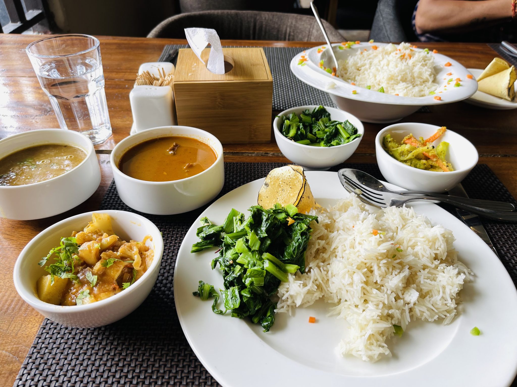 Nepali Lunch/Food. Large plate on the right with rice and some greens on it, three small bowls to the left with soups or gravies, tissues, toothpicks and a glass of water in the center of the table.
