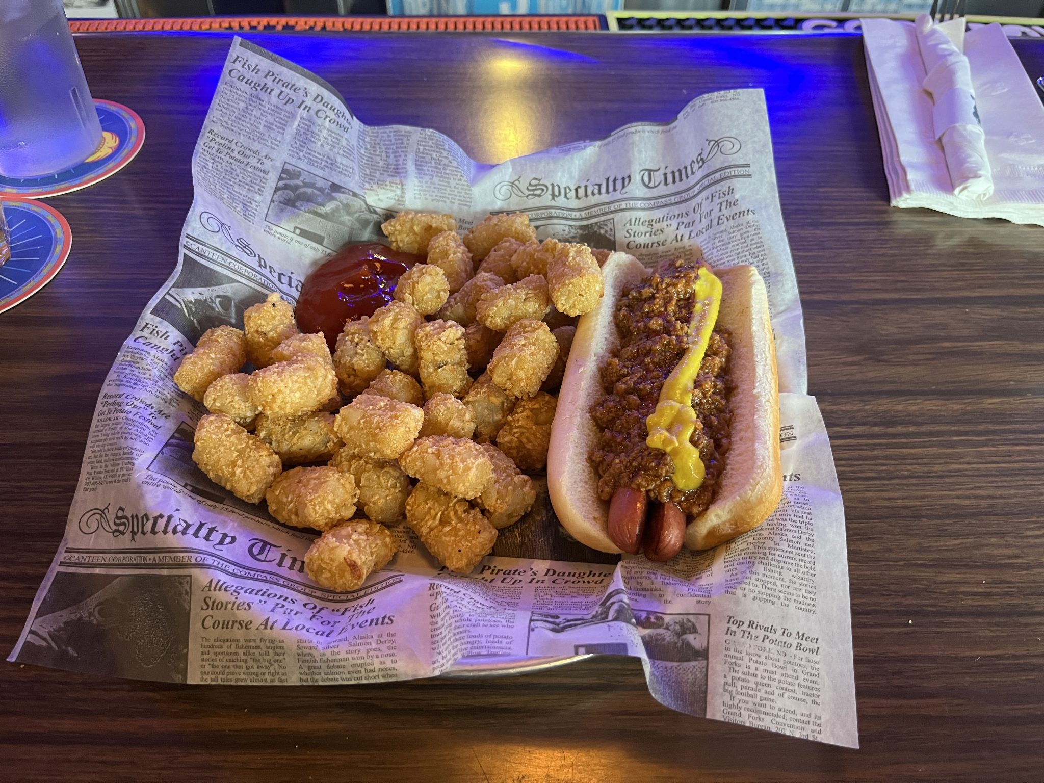 A plastic bowl lined with fake newsprint paper holding a chilidog (on the right) with a stripe of mustard and a big pile of tater tots on the left with a puddle of catsup next to them.