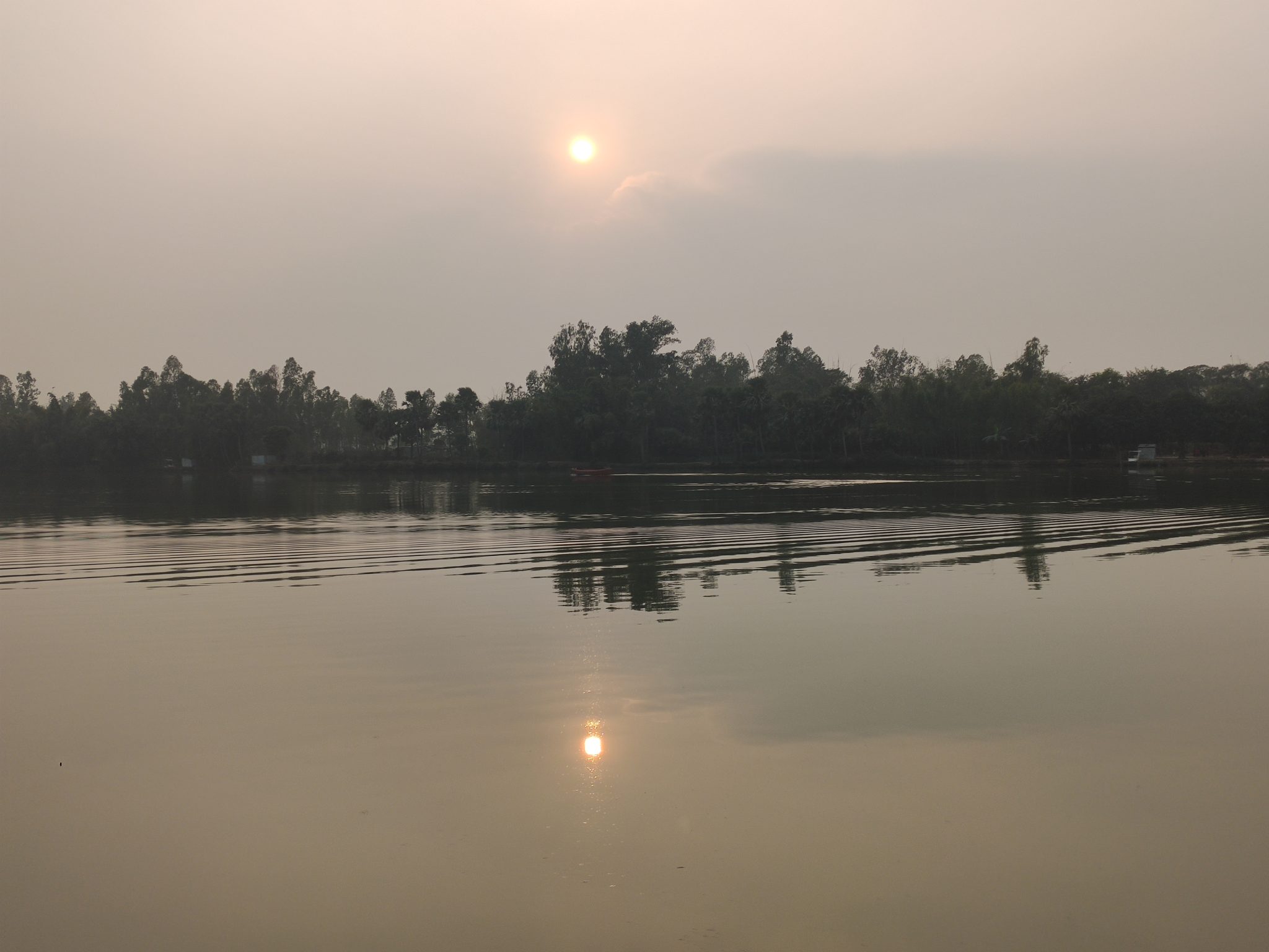 A calm lake or river in the foreground, with forest on the far side.  The sun is just over the horizon but mostly hidden behind clouds so the world is dim.