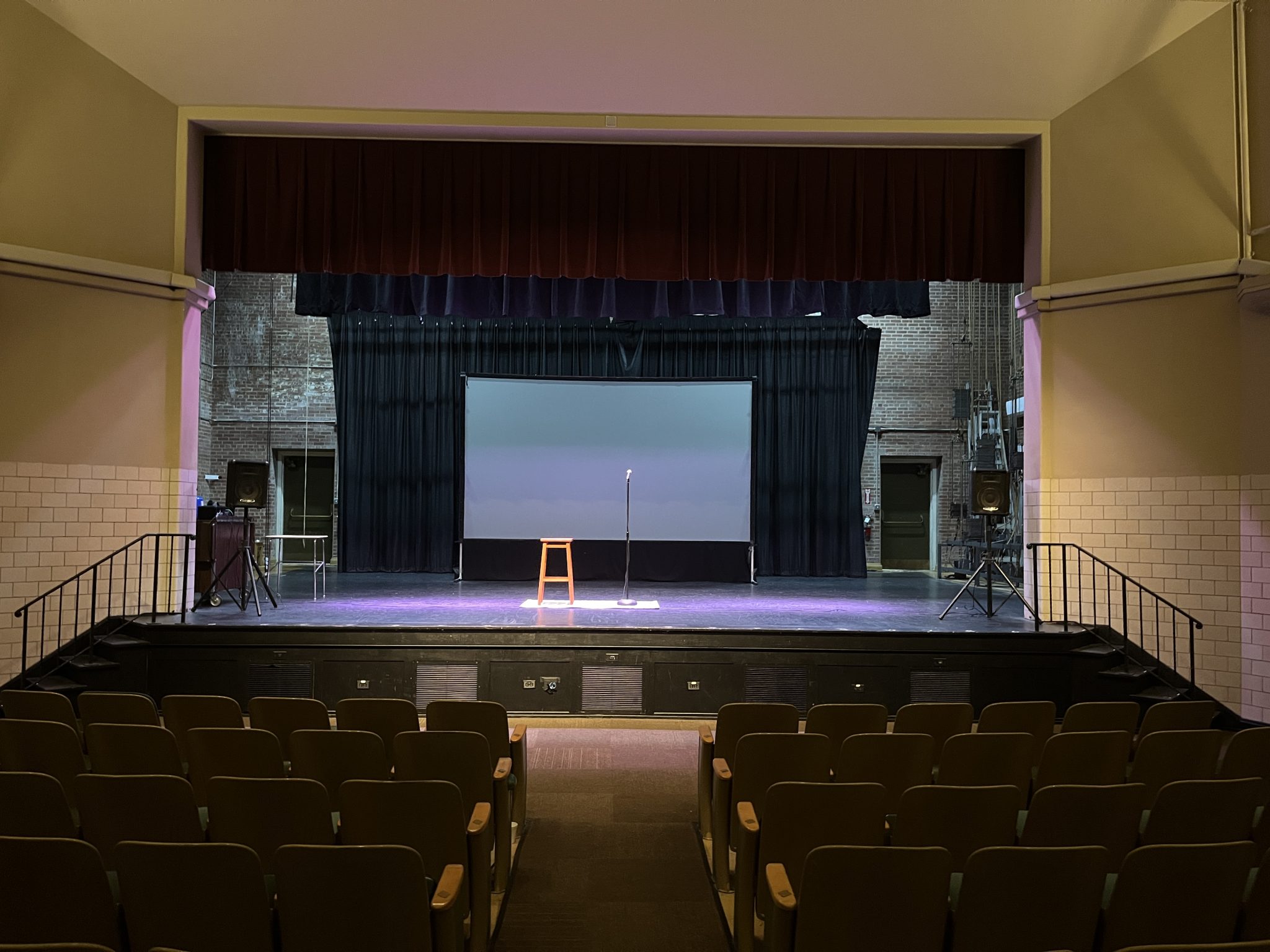 A stage lit from above with nothing on it except a small carpet, a stool, and a microphone. Perspective is from the back of the auditorium, so you can see many empty chairs in the foreground.