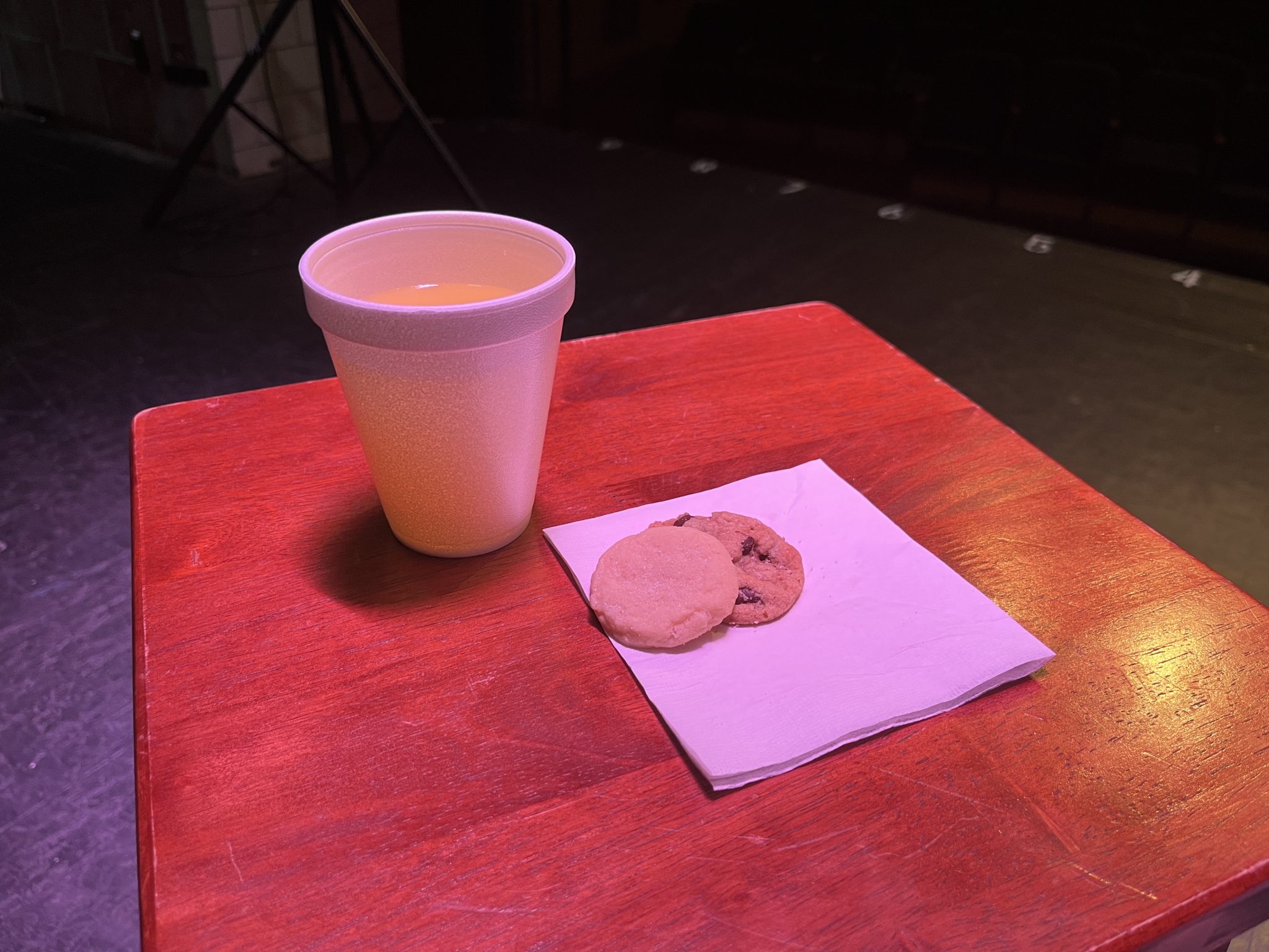 two cookies on a napkin on a small table, with a cup of lemonade, under a spotlight.