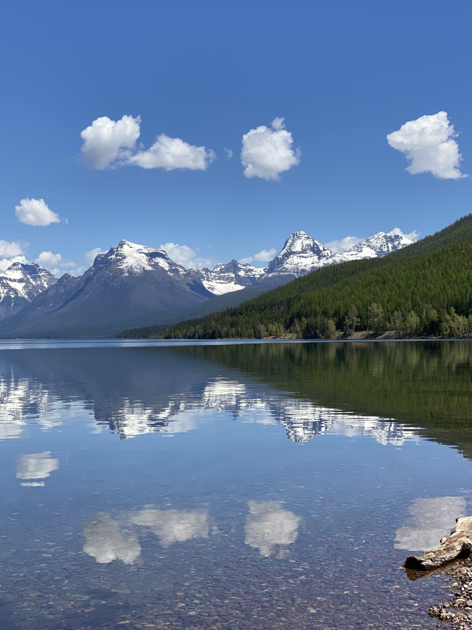 Lake McDonald in Glacier National Park.  Water in the immediate foreground, reflecting the snowy mountain in the distance. Small puffy clouds in an otherwise clear sky.