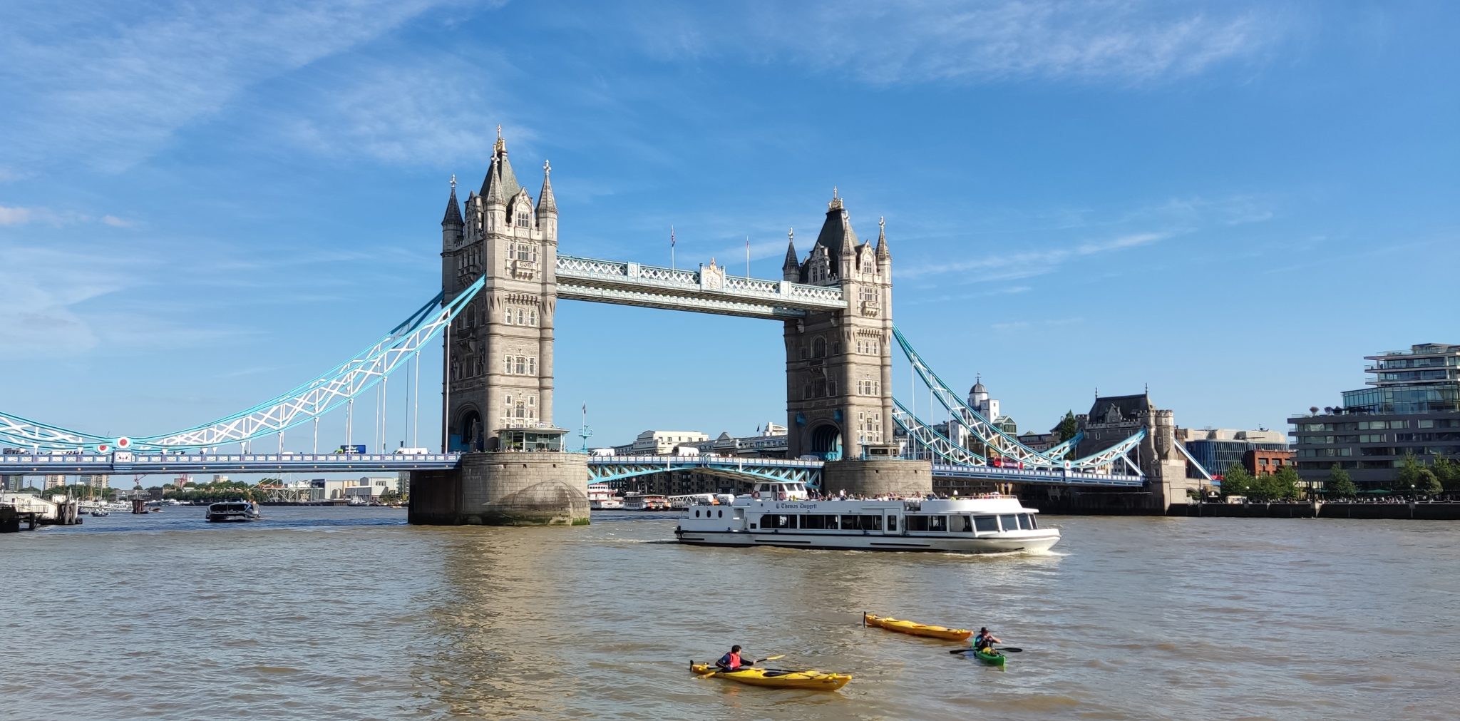 Tower Bridge, London. Photographer is on the water, it's a sunny day, and in the foreground is a large tour boat and two single-person kayaks.
