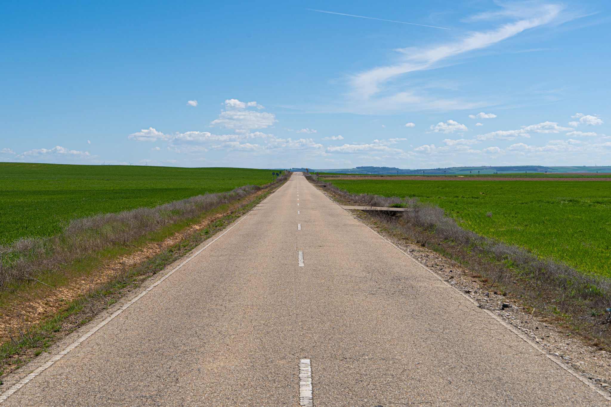 Photo of a lonely road that disappears into the horizon and crosses a field of green grass with blue sky and a few clouds.