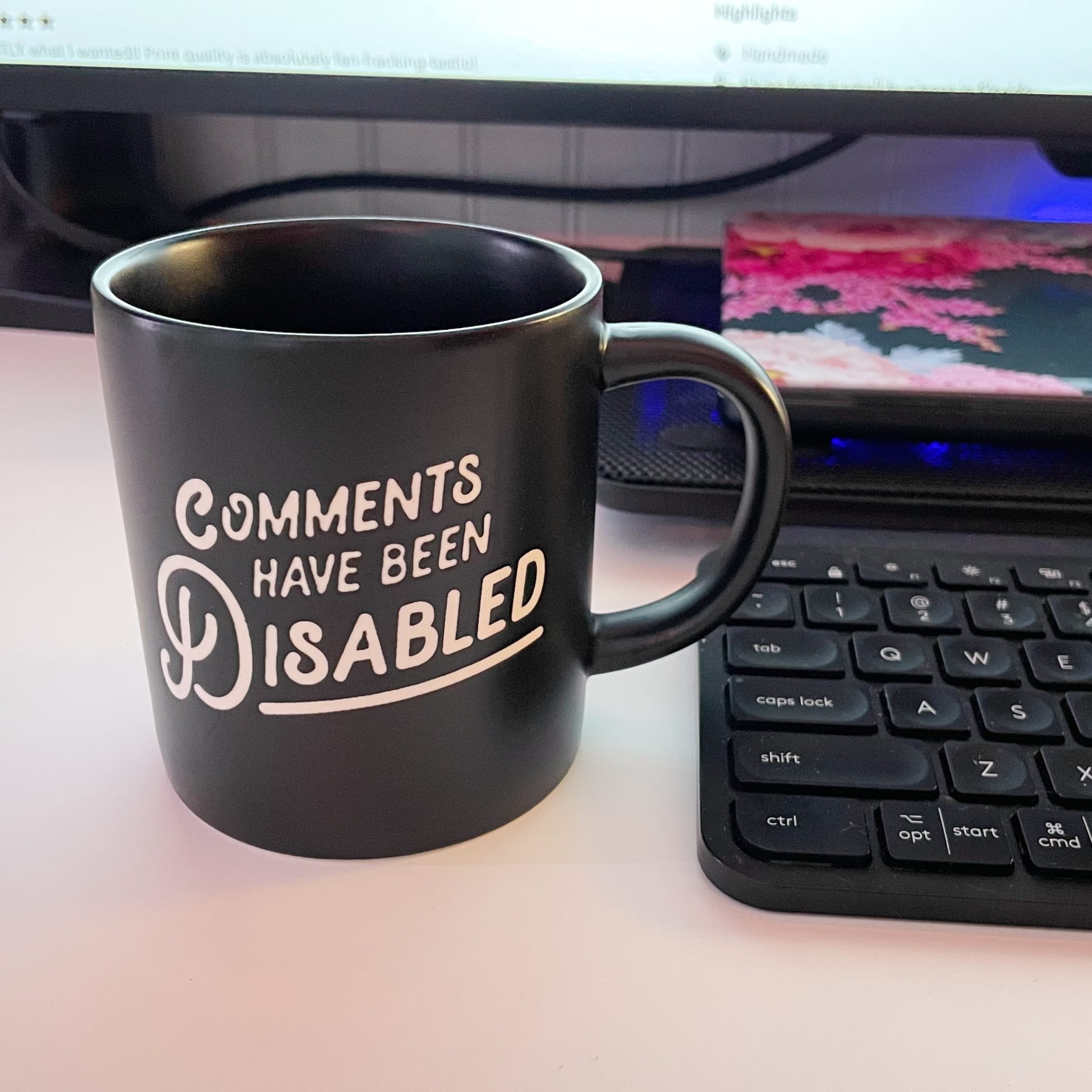A mug with the text, "Comments Have Been Disabled" on it, next to a keyboard and a laptop on a cooling mat behind it
