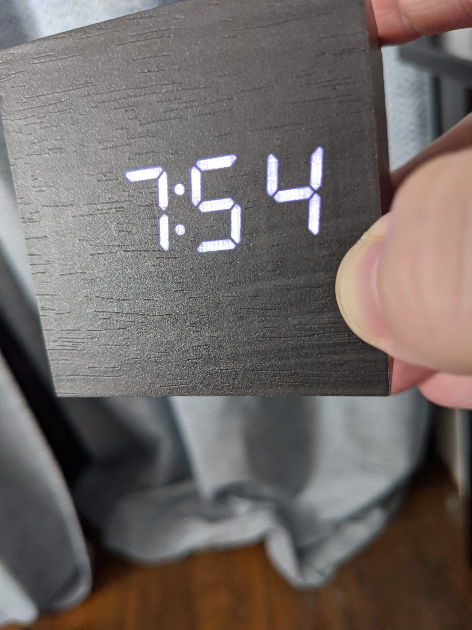 A square digital clock, held in someone's hand. It is 7:54